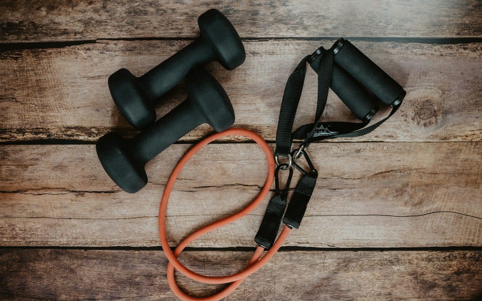 Two black dumbbells and an orange resistance band on a rustic wooden surface
